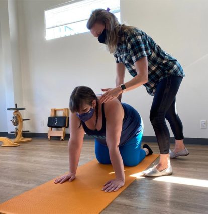 woman on yoga mat on hands and knees while another woman pushes down on back and shoulder