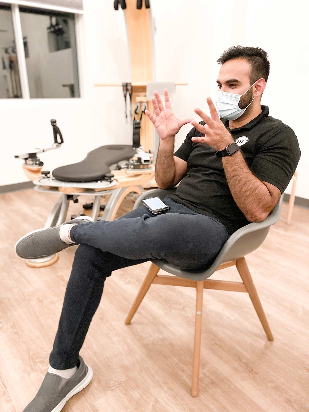 man wearing a surgical mask sitting in a chair with his left foot over his right knee and gesturing