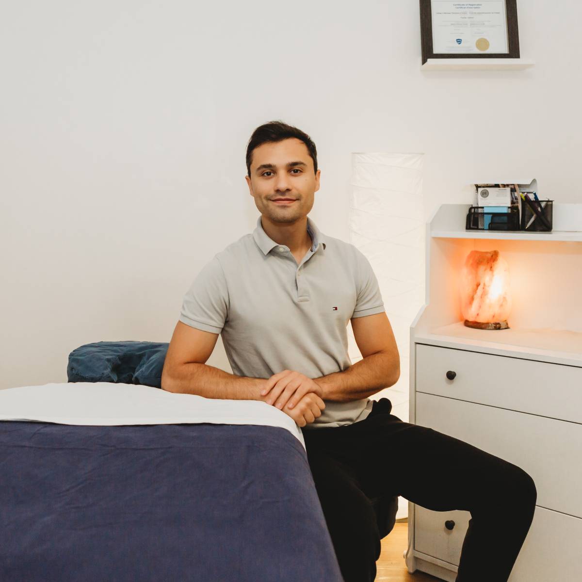 Physical therapist seated beside a treatment table in a clean clinic setting, with a framed certificate and softly lit salt lamp visible in the background.