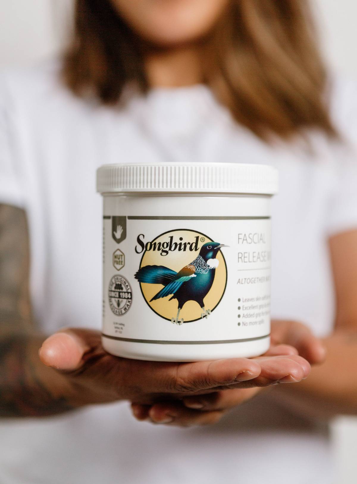 Person holding a container of Songbird Fascial Release Cream in their hands, with the product label prominently displayed against a softly blurred background.