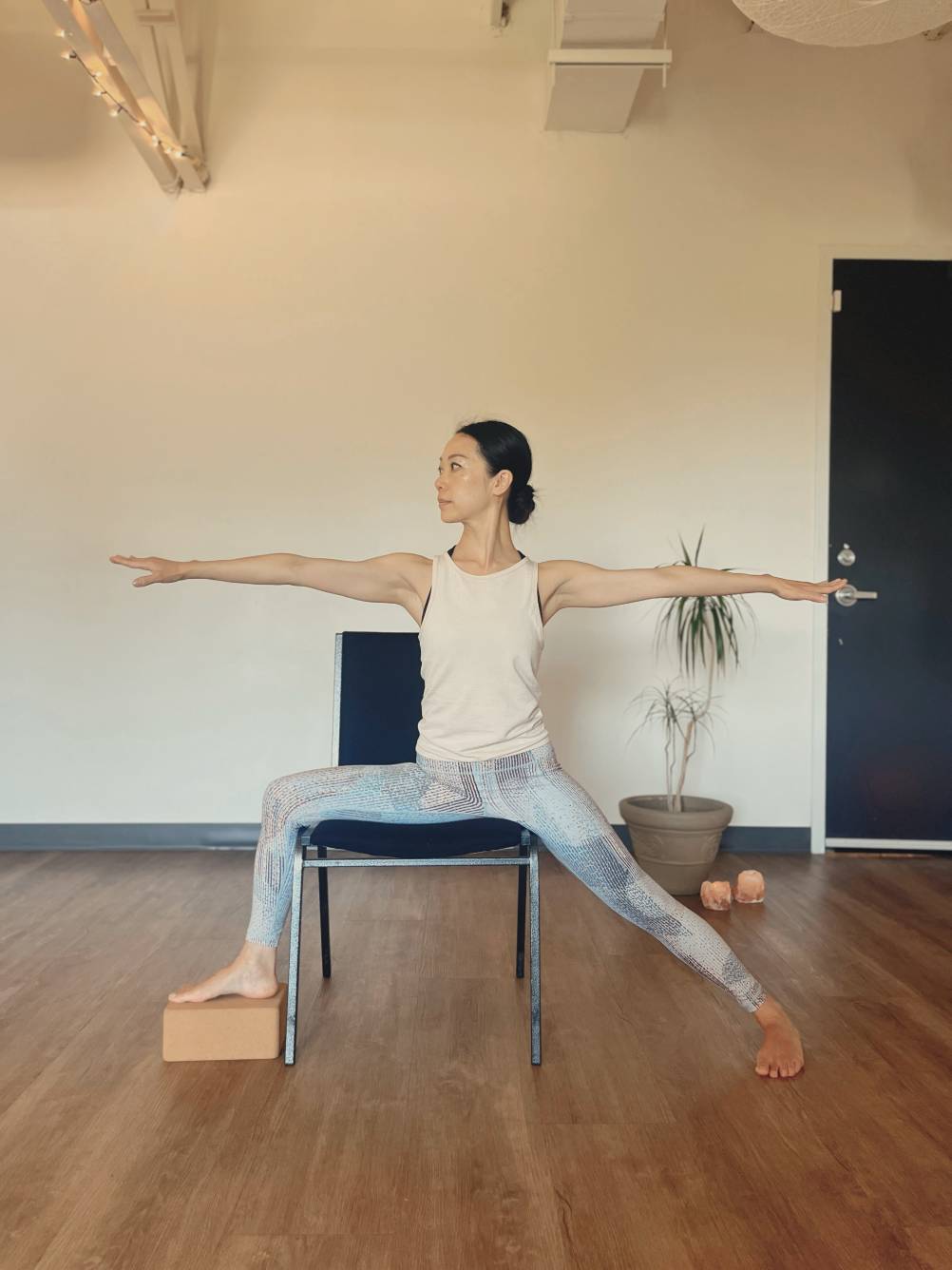 Woman practicing seated yoga in a chair with arms extended and one foot supported by a yoga block.